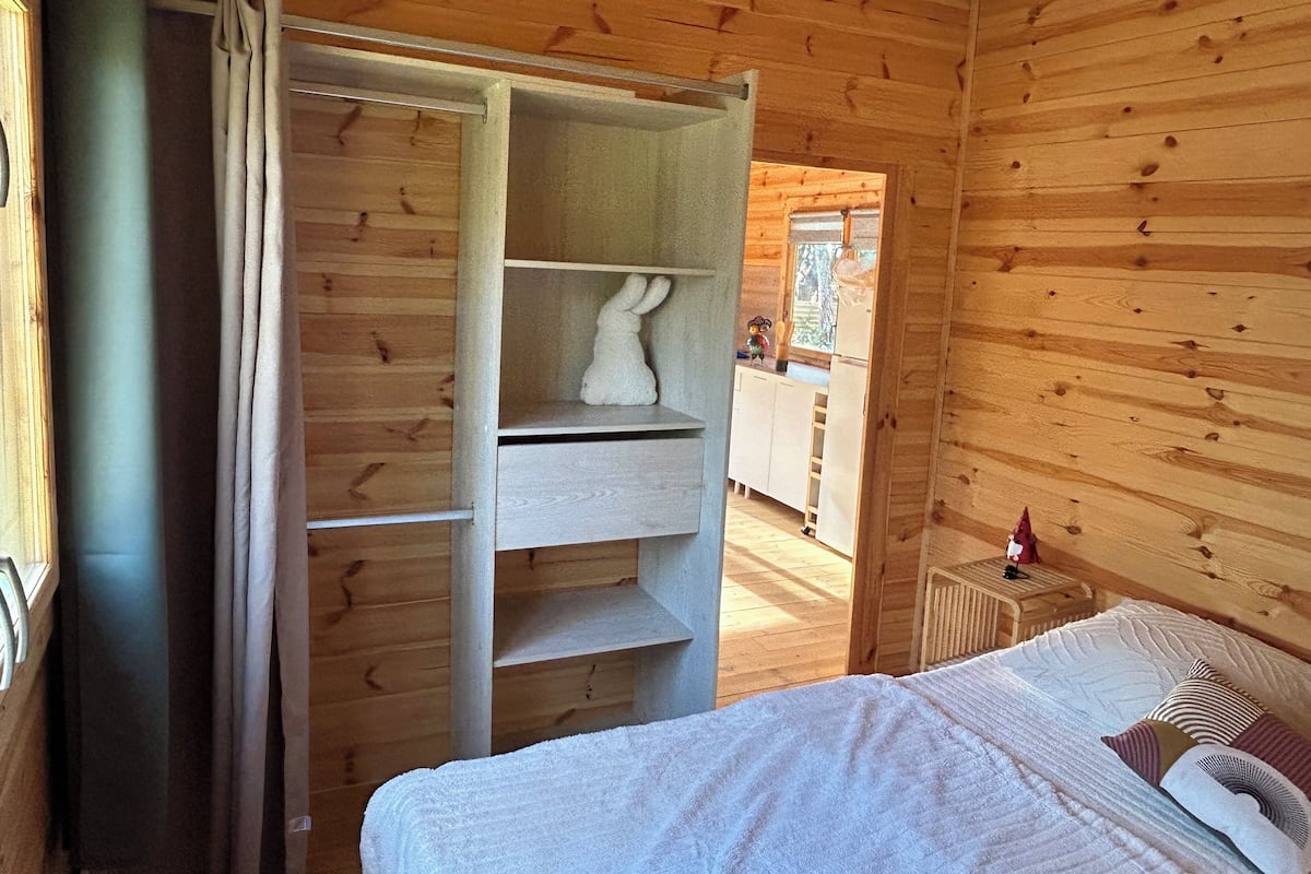 A wooden cabin bedroom with a bed, a white towel, a shelf with a white object, and a window with a view of the outside.