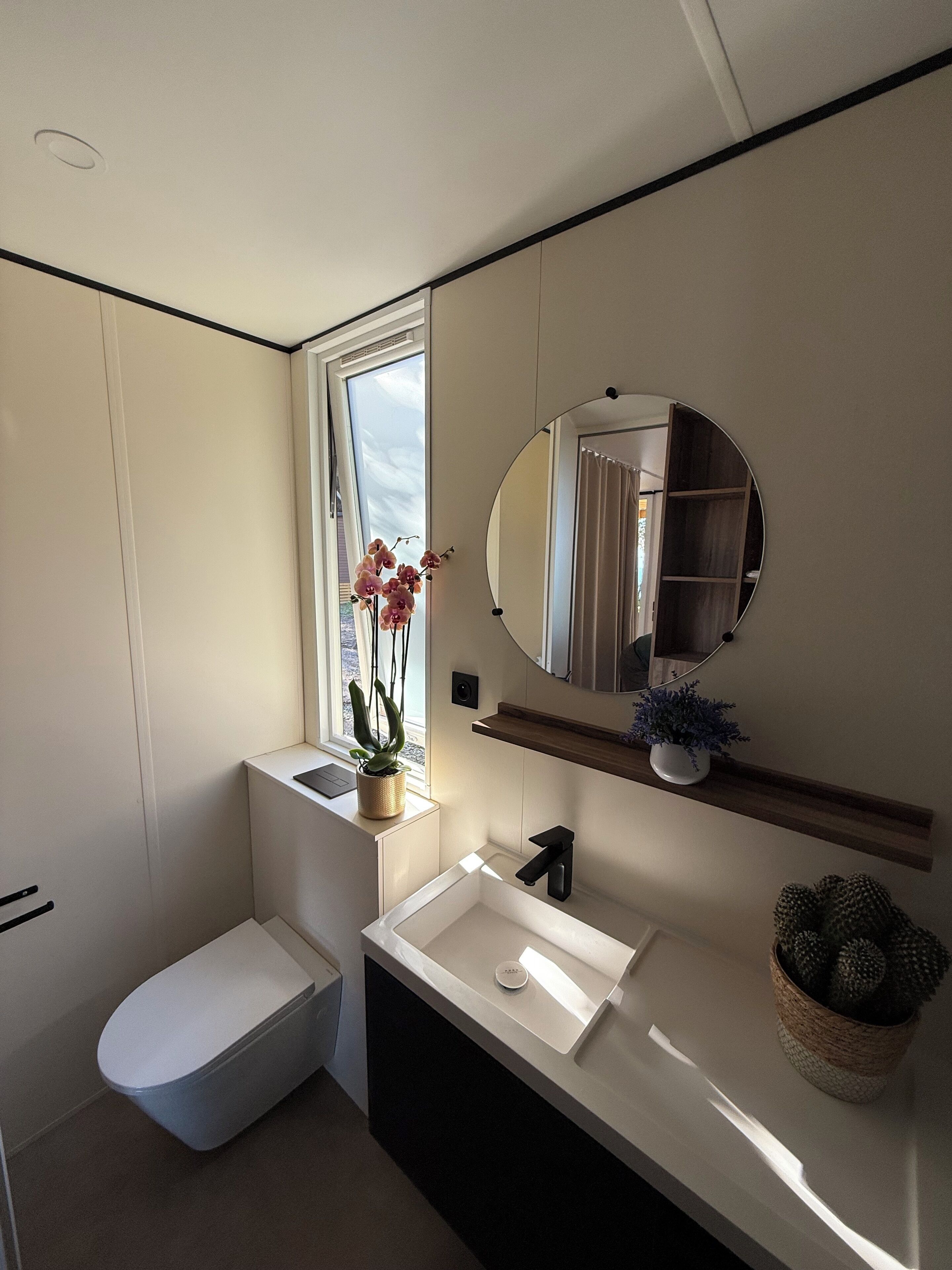 Ein modernes Badezimmer mit Toilette in Weiß, einem Waschbecken mit Spiegel darüber und einem Fenster mit Orchideen.