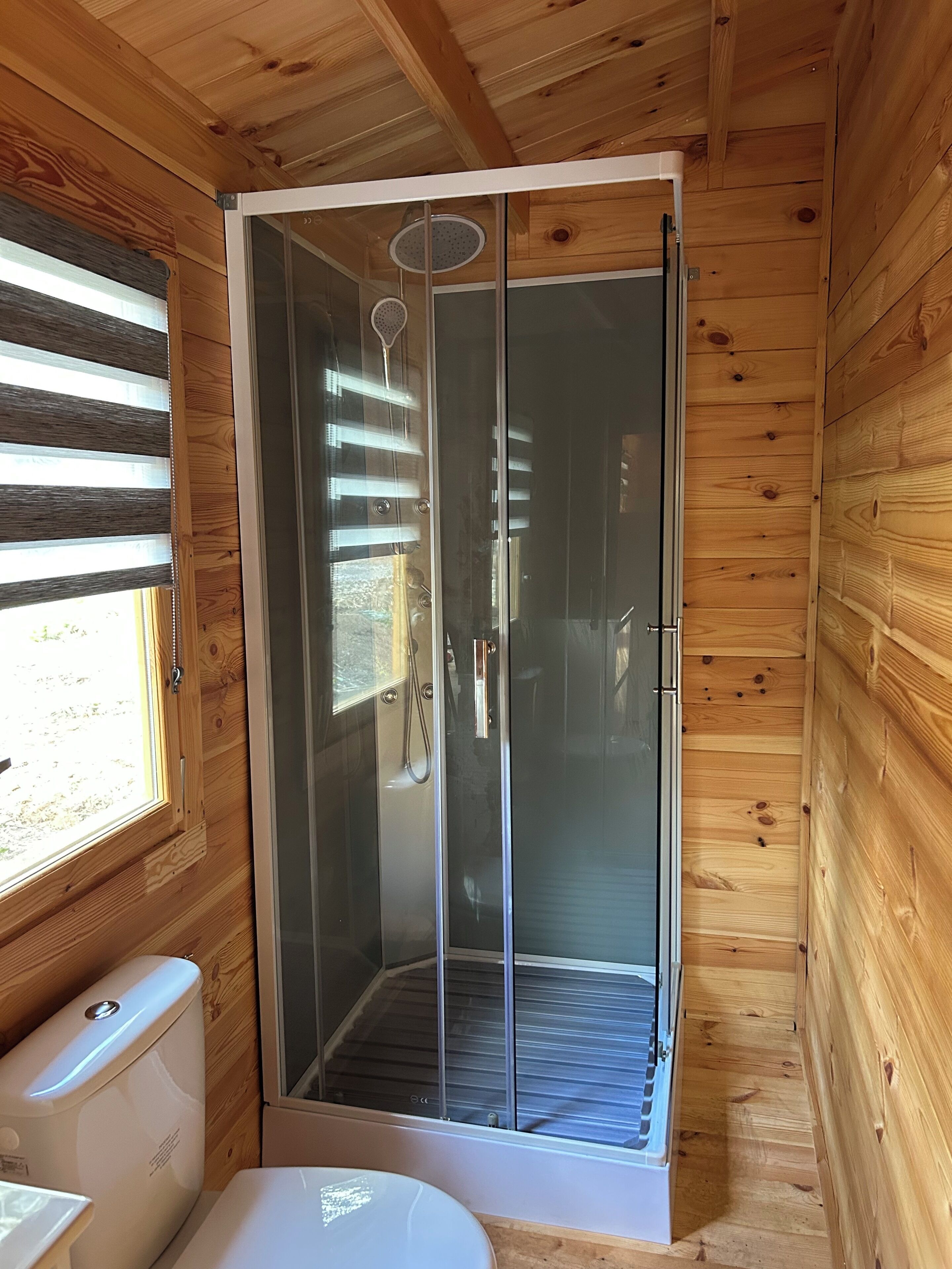 Un baño de cabaña de madera con ducha envuelta en vidrio, un inodoro y una ventana con persianas.