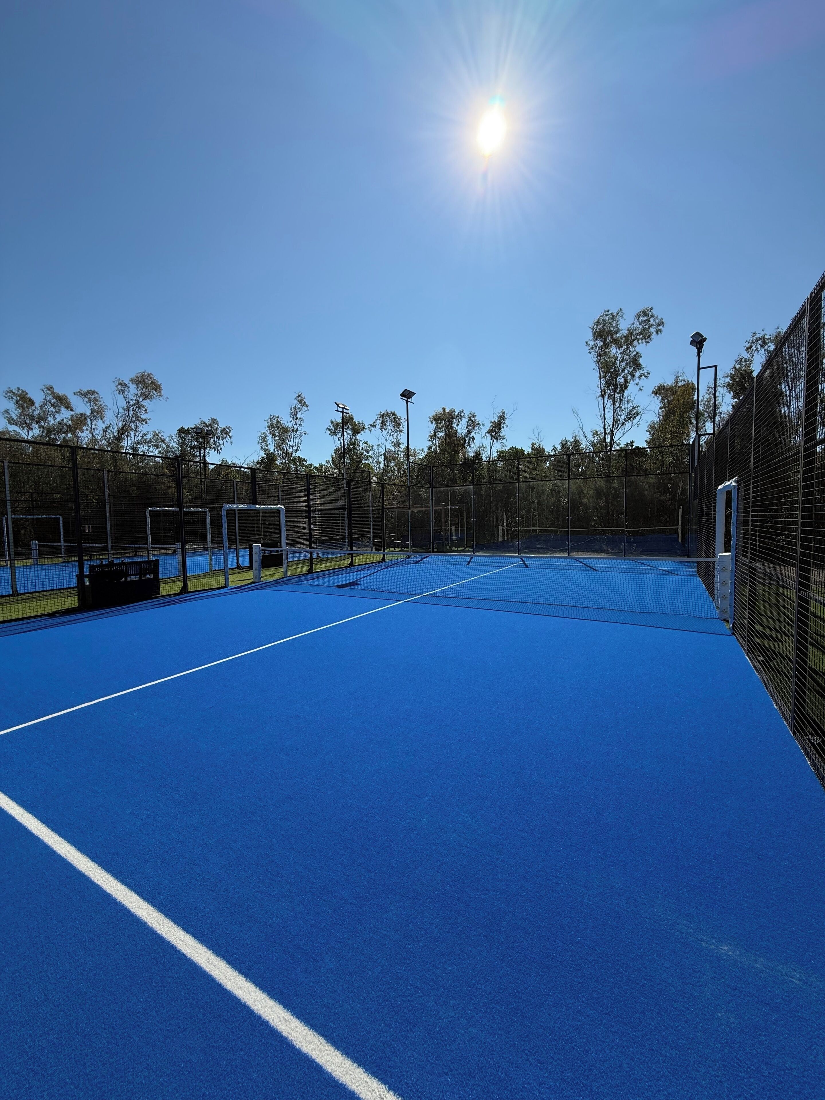 Een buitenbaan voor tennis met een blauwe ondergrond, omringd door een zwarte hekken, en bomen op de achtergrond.