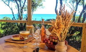 A table set with a bottle, glasses, a bowl of grapes, and a plate of fruit, overlooking a coastal view. - Calella Camping(Naturiste) (Linguizzetta)