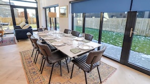 A dining area with a large table set for six, surrounded by chairs, with a view of a fenced yard through large windows. - The Barn (Morpeth)