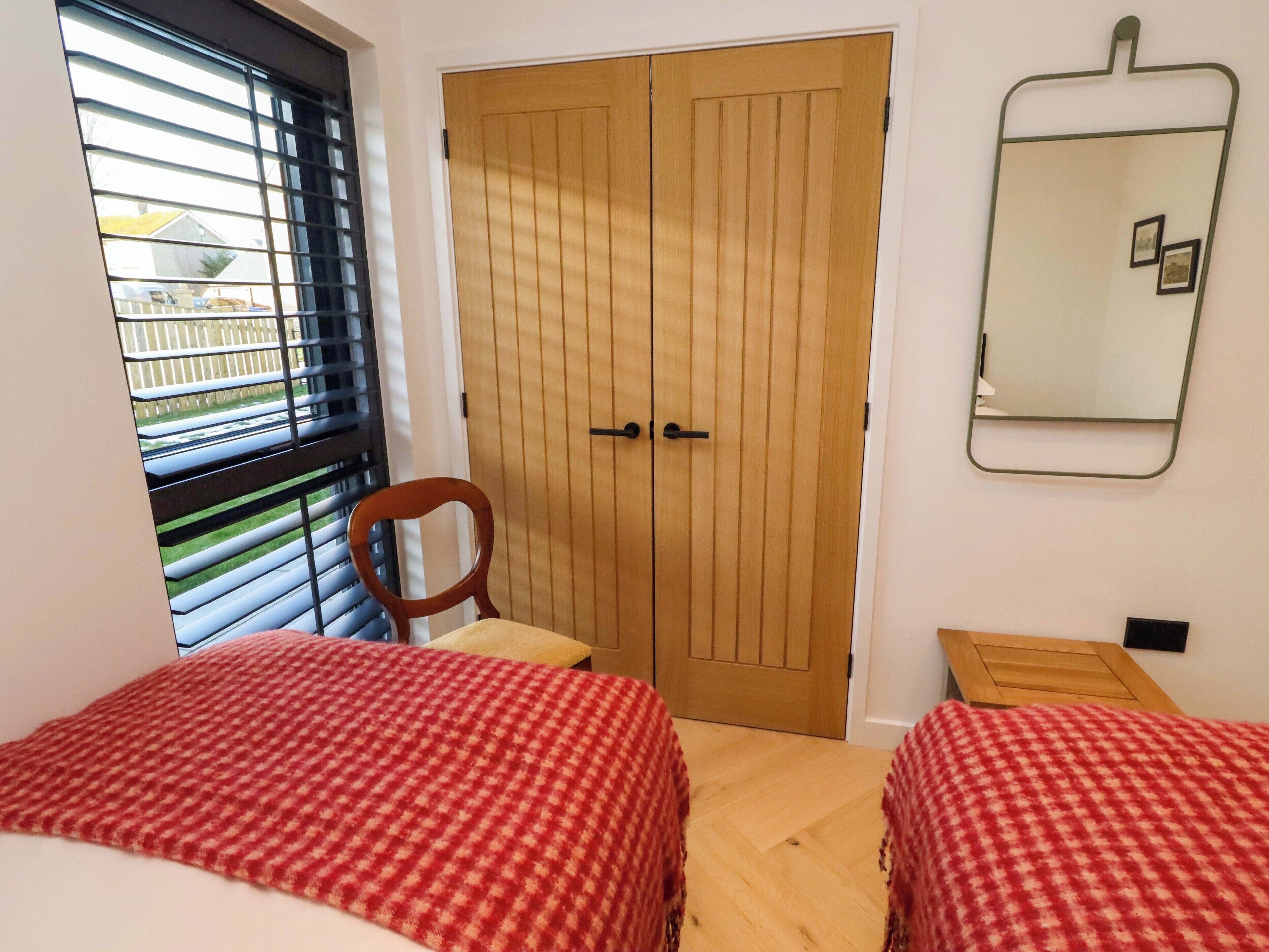A bedroom with two beds, a wooden wardrobe, a mirror, and a window with blinds.