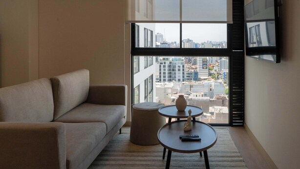 A modern living room with a sofa, a round table with a vase and remote, and a large window with blinds. - w* | Amazing 1BR w/ Balcony in Barranco (Barranco)