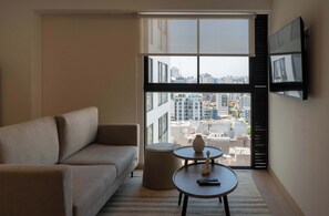 A modern living room with a sofa, a round table with a vase and remote, and a large window with blinds. - w* | Amazing 1BR w/ Balcony in Barranco (Barranco)