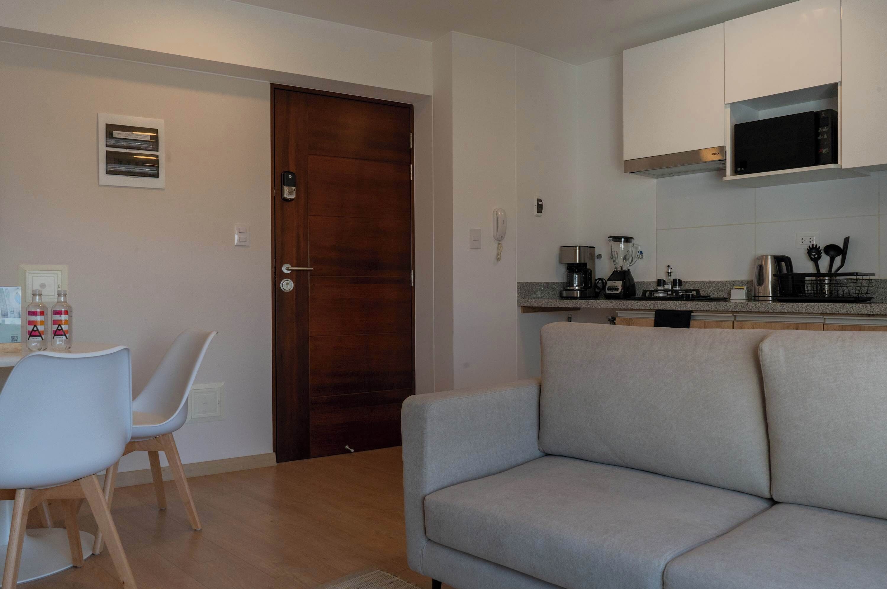 Une cuisine moderne avec des armoires blanches, un four à micro-ondes encastré et une machine à café. On y trouve une porte en bois et un canapé beige dans l’espace salon.