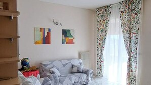 Ein Wohnzimmer mit einer Sofagarnitur, einem Couchtisch und einer Heizung. An der Wand befinden sich zwei abstrakte Gemälde und ein Fenster mit floralen Vorhängen.