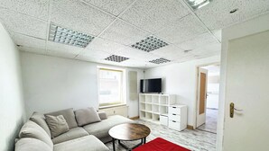 Een moderne woonkamer met een hoekbank, een klein rond tafeltje, een rode poef, een televisie op een witte dressoir, en een raam met jaloezieën.