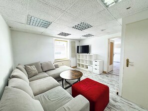 A modern living room with a sectional sofa, a small round table, a red ottoman, a TV on a white stand, and a window with blinds. - Central Berlin Apartment for 8 with Parking (Berlin)