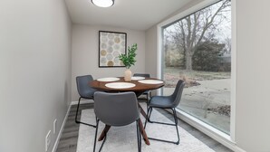 Ein moderner Essbereich mit einem runden Tisch, vier Stühlen, einer Pflanze in einer Vase und einem großen Fenster mit Blick auf Bäume und eine Auffahrt.