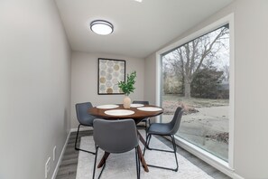 A modern dining area with a round table, four chairs, a vase with a plant, and a large window offering a view of trees and a driveway. - Modern East Side Ranch (Wichita)