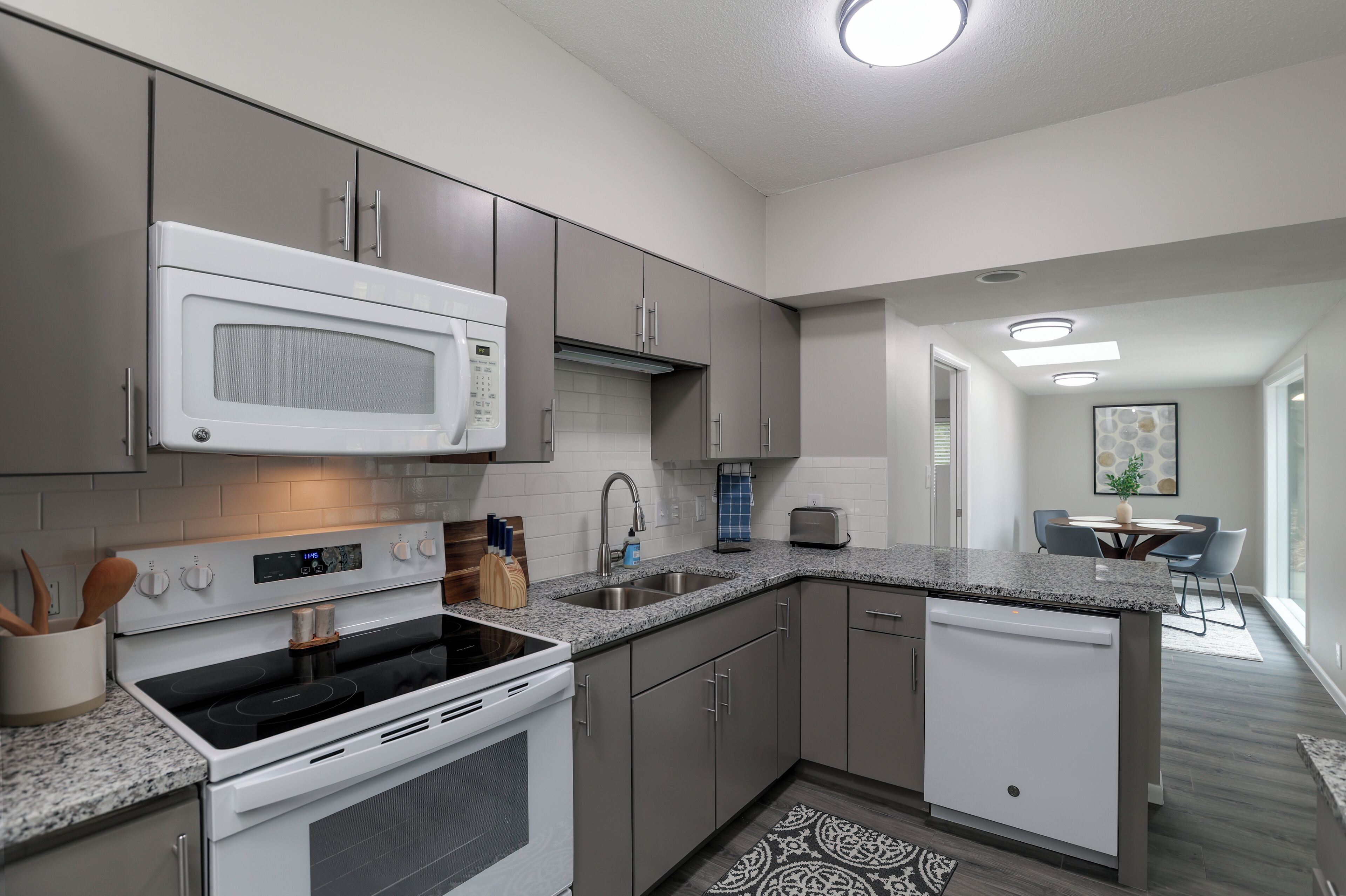 A modern kitchen with a microwave, stove, sink, and dishwasher. There is a dining area with a table and chairs visible in the background.