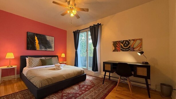 Quarto com cama, escrivaninha, ventoinha no teto e janela com cortinas.