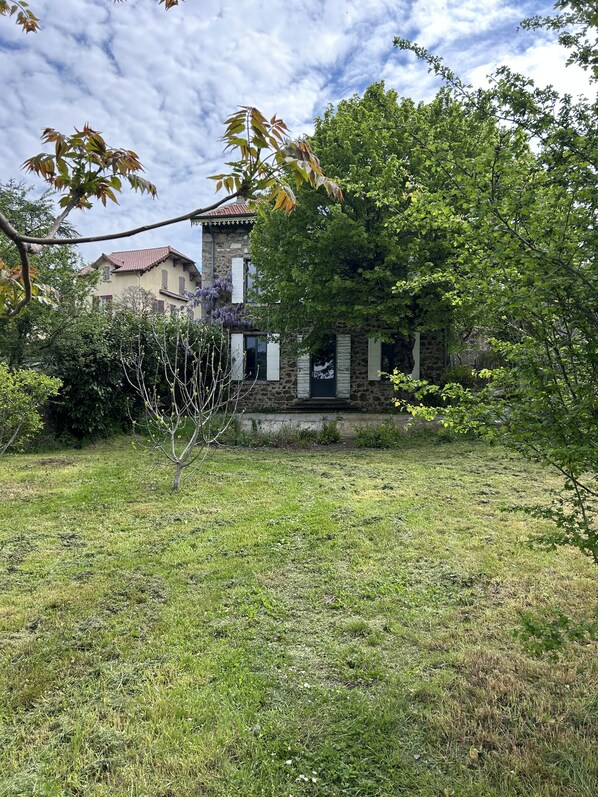 Un bâtiment en pierre, surmonté d’un toit en tuiles, entouré d’une zone herbeuse agrémentée d’arbres et d’arbustes.