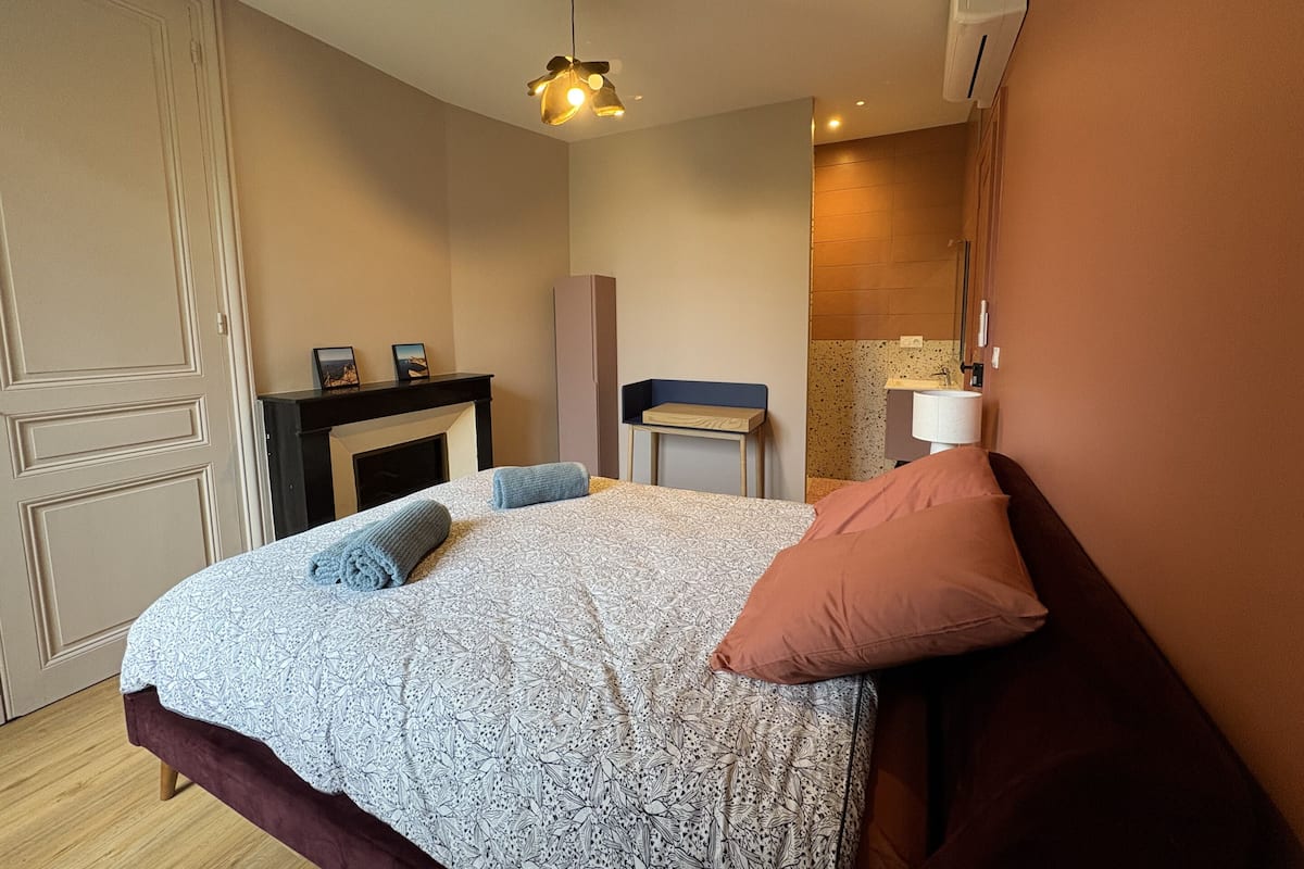 A bedroom with a bed, a bedside table, a mirror, a fireplace, and an air conditioning unit.