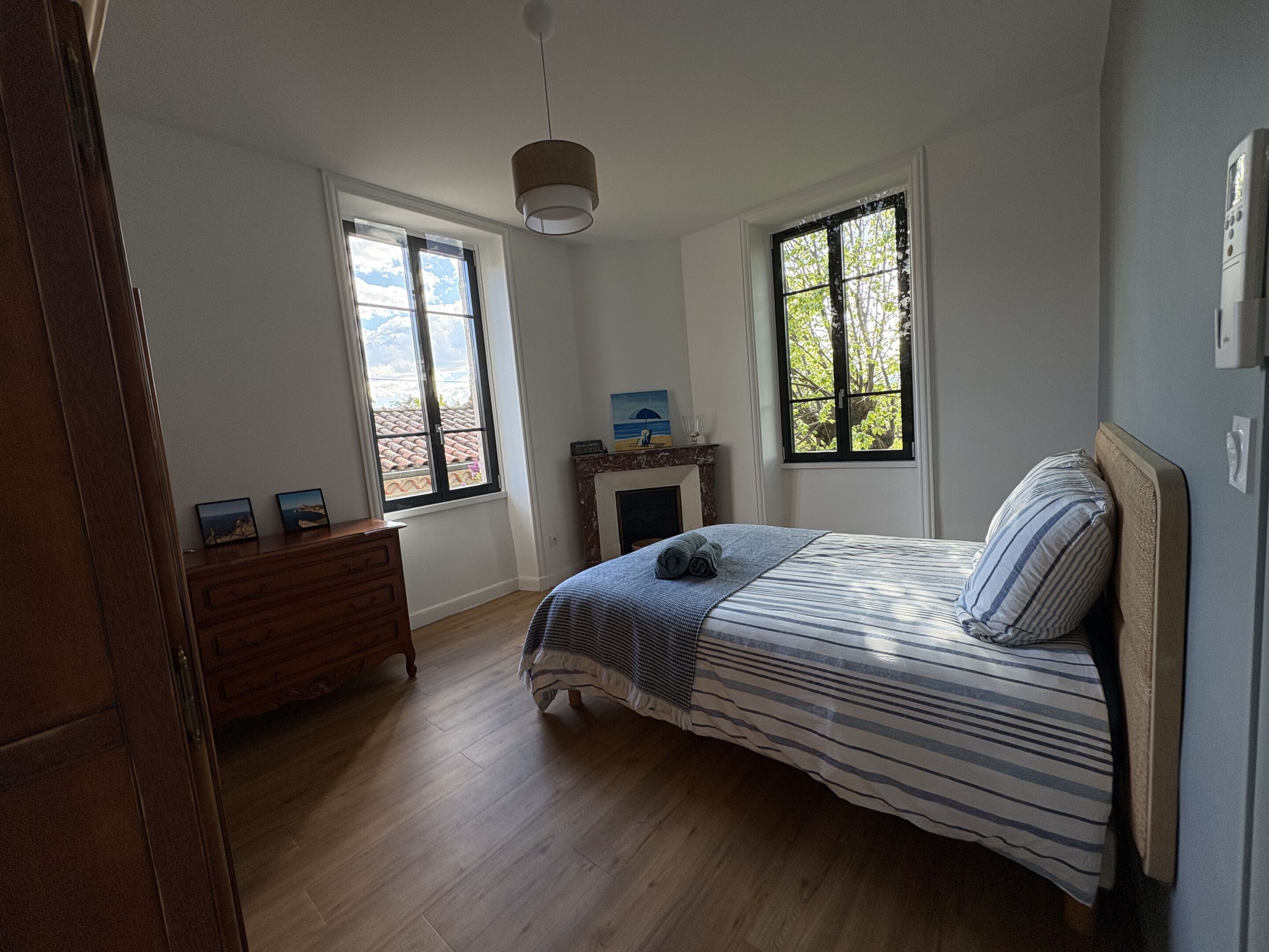 Un dormitorio con cama, dos ventanas, un aparador y una chimenea.