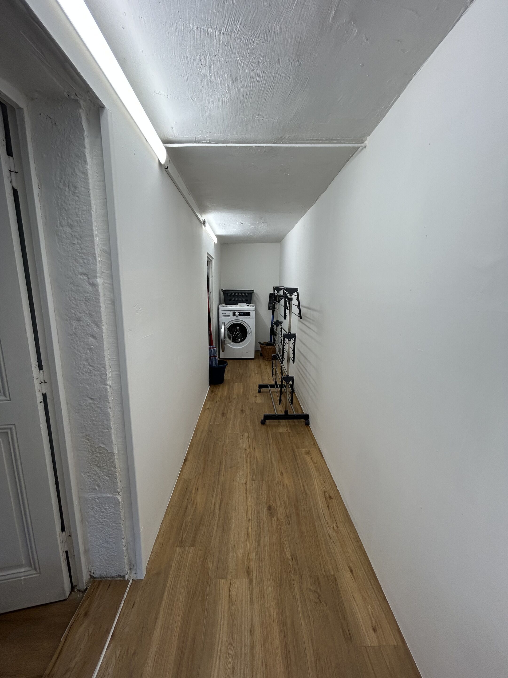 Un couloir étroit avec un plancher en bois, des murs blancs et un luminaire au plafond. On aperçoit une machine à laver et un étendoir au loin.