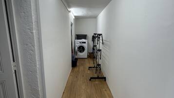 A narrow hallway with wooden flooring, white walls, and a ceiling light. A washing machine and a drying rack are visible in the distance.