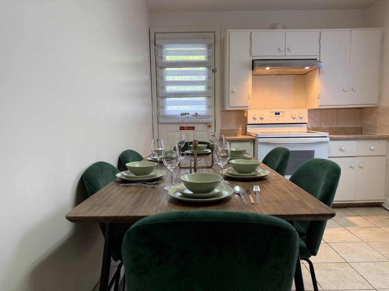 Un comedor con una mesa de madera dispuesta para cuatro personas, sillas verdes alrededor y una cocina al fondo.