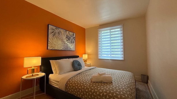 Ein Hotelzimmer mit Bett, Nachttisch, Lampe, einem Fenster mit Jalousien und einem Kunstwerk an der Wand.
