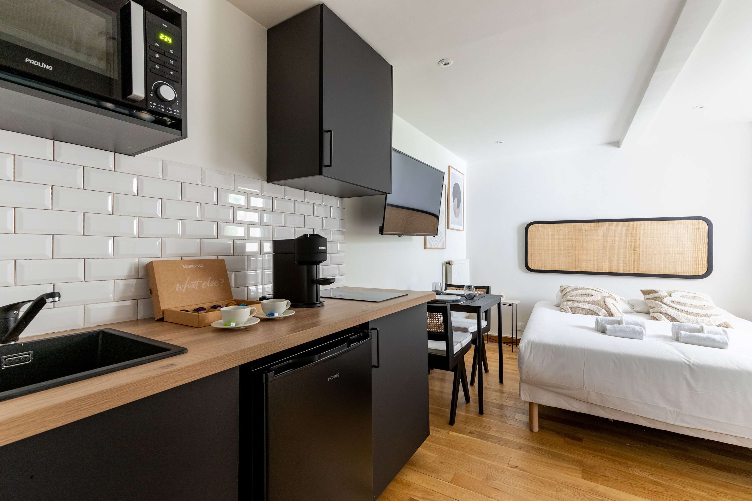 Cocina compacta con microondas, cafetera y lavavajillas. Un pequeño comedor con mesa y sillas, y una cama con ropa de cama blanca en la habitación contigua.
