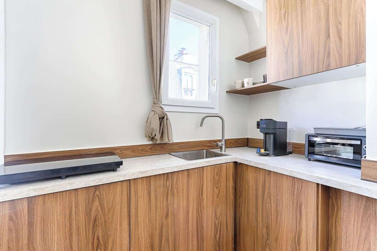 A modern kitchen with wooden cabinets, a built-in oven, a coffee maker, and a window with a view of a building.