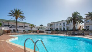 Une piscine avec une terrasse carrelée, des palmiers et un bâtiment doté de balcons en arrière-plan.