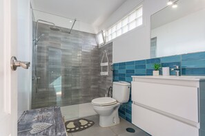 Kylpyhuoneessa on lasiseinäinen suihkukaappi, wc-istuin, peilillä varustettu pesuallas ja pieni kasvi tasolla.