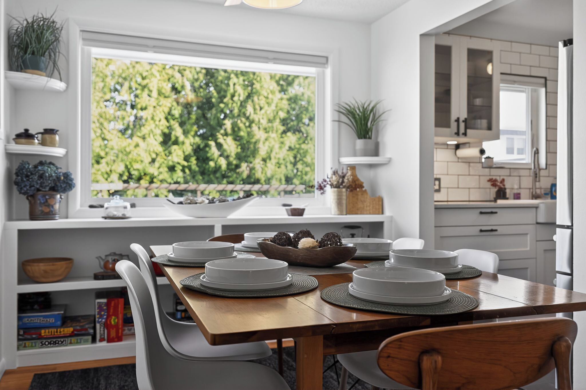 A dining area with a wooden table set for four, white plates, and a bowl of fruit. There are chairs, a window with a view of greenery, and a kitchen in the background.