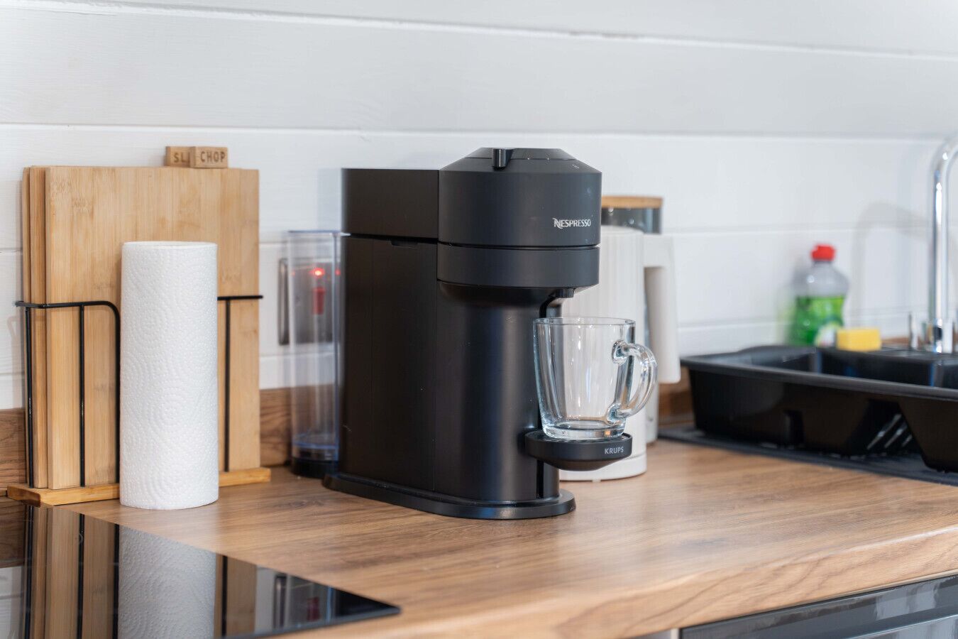 Een moderne keuken met een zwarte Nespresso koffiemachine, een glazen mok en een houten snijplank met keukenpapier.