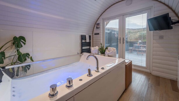 A modern bathroom with a large bathtub, a sink, and a shower. There is a plant, towels, and a television mounted on the wall. - Apex Studio Pod & Jacuzzi Bath (Fort William)