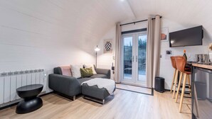 A modern, well-lit room with a grey sofa, a small round table, and a kitchen area with stools. - Apex Studio Pod & Jacuzzi Bath (Fort William)