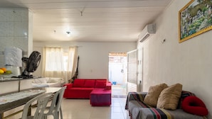Uma sala de estar com um sofá vermelho, um sofá listrado, uma mesa de jantar com cadeiras e uma área de cozinha com um ventilador e uma geladeira.