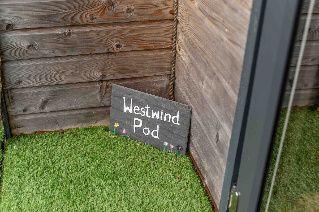 A wooden wall with a chalkboard sign reading "Westwind Pod" on a grassy surface.