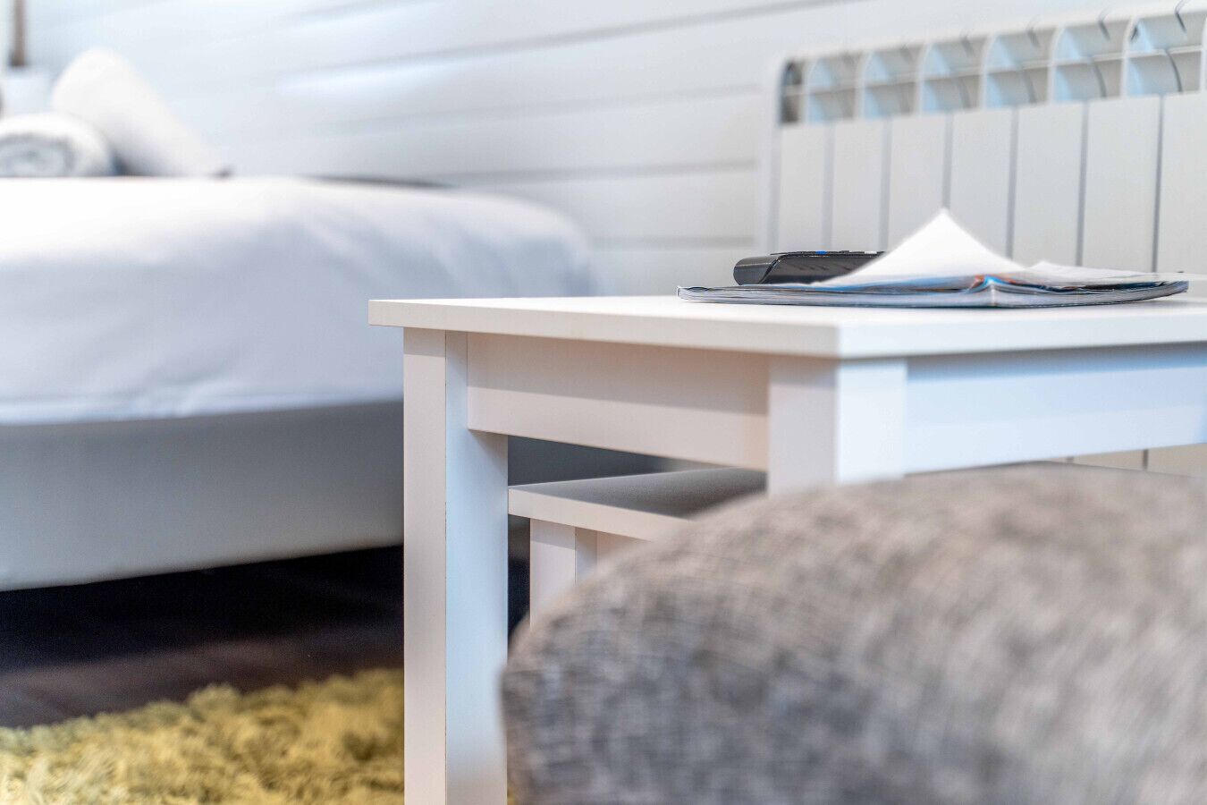 Una mesita de noche blanca con un teléfono inteligente y un libro en un dormitorio moderno con cama blanca y cabecera a rayas.