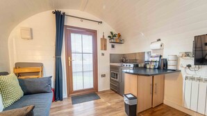 Une cuisine compacte équipée d’une cuisinière, d’un four et d’un plan de travail, un petit coin salon avec un canapé et un fauteuil, et une porte donnant sur l’extérieur.