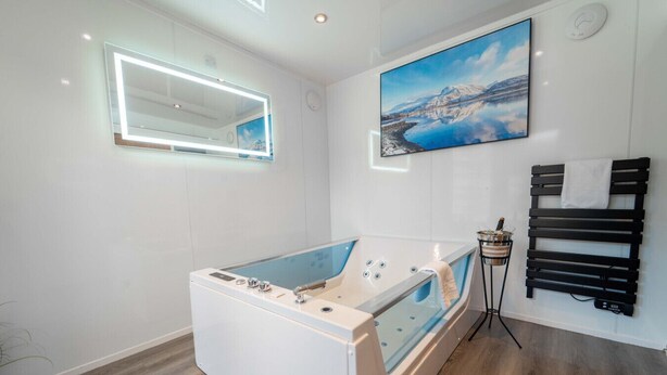 A modern bathroom with a freestanding bathtub, a large mirror with integrated lighting, a painting of a mountainous landscape, and a black towel rack. - Ben View Room & Jacuzzi Bath (Fort William)