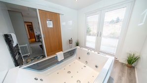A modern bathroom with a large bathtub, a wooden door, a small plant, and a window with blinds. - Ben View Room & Jacuzzi Bath (Fort William)