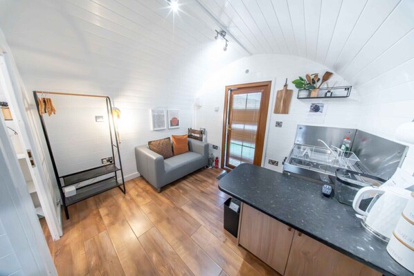 A compact living space with a kitchenette, a grey sofa, wooden flooring, and a ceiling with exposed beams. - Ben View Pod & Jacuzzi Bath (Fort William)