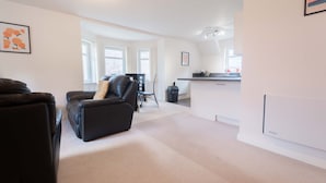 A spacious living room with a black leather sofa, a dining area with a table and chairs, and a kitchen area with a countertop and cabinets.