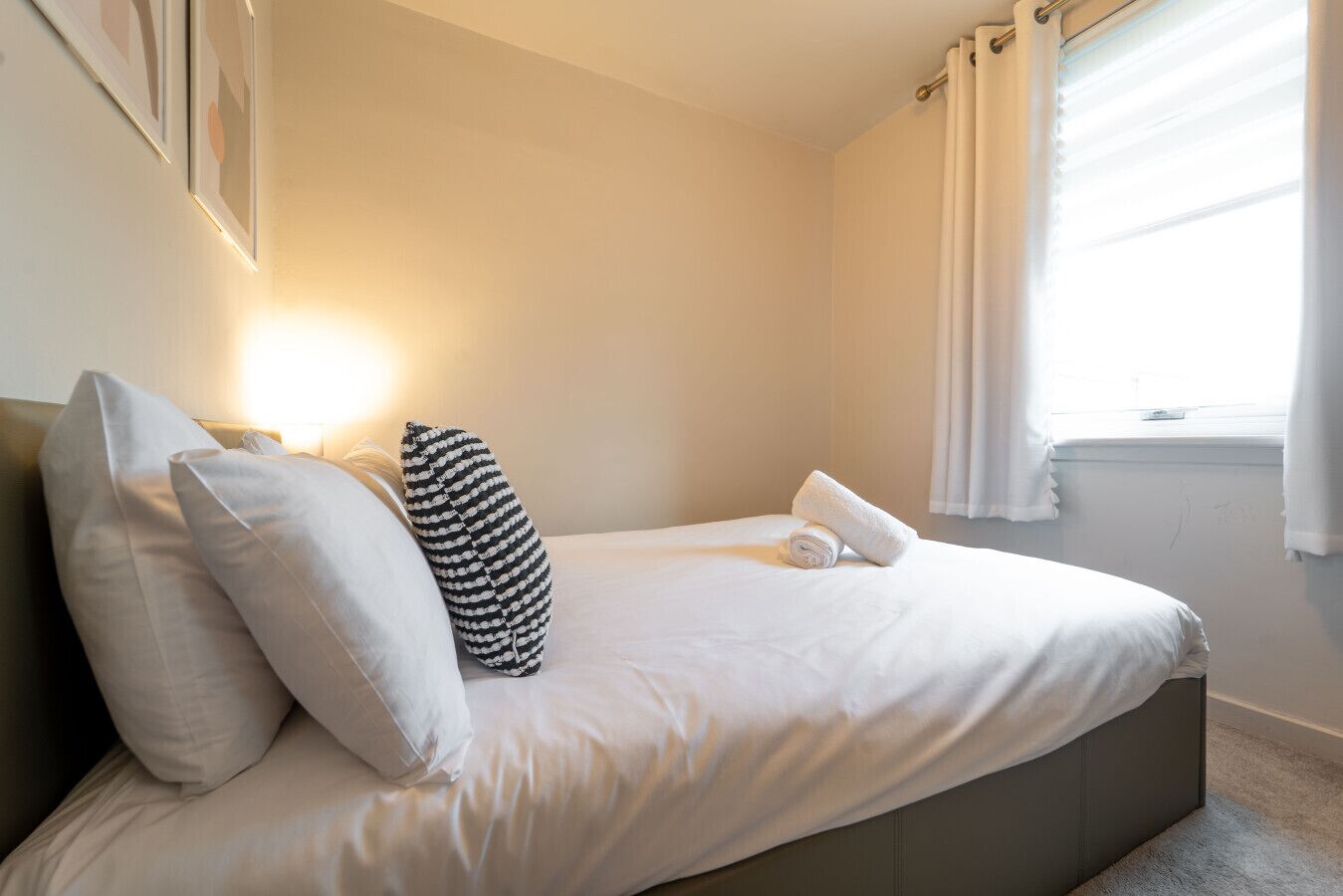 A neatly made bed with white linens, a striped pillow, and a rolled towel.