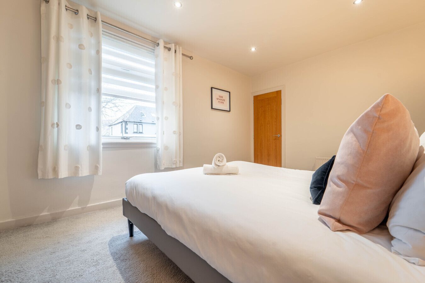A bedroom with a bed, a window with curtains, a door, and a framed picture on the wall.
