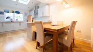 Een moderne keuken met witte kasten, een houten eettafel en twee stoelen. Boven de keuken zijn dakramen en erboven hangen hanglampen.