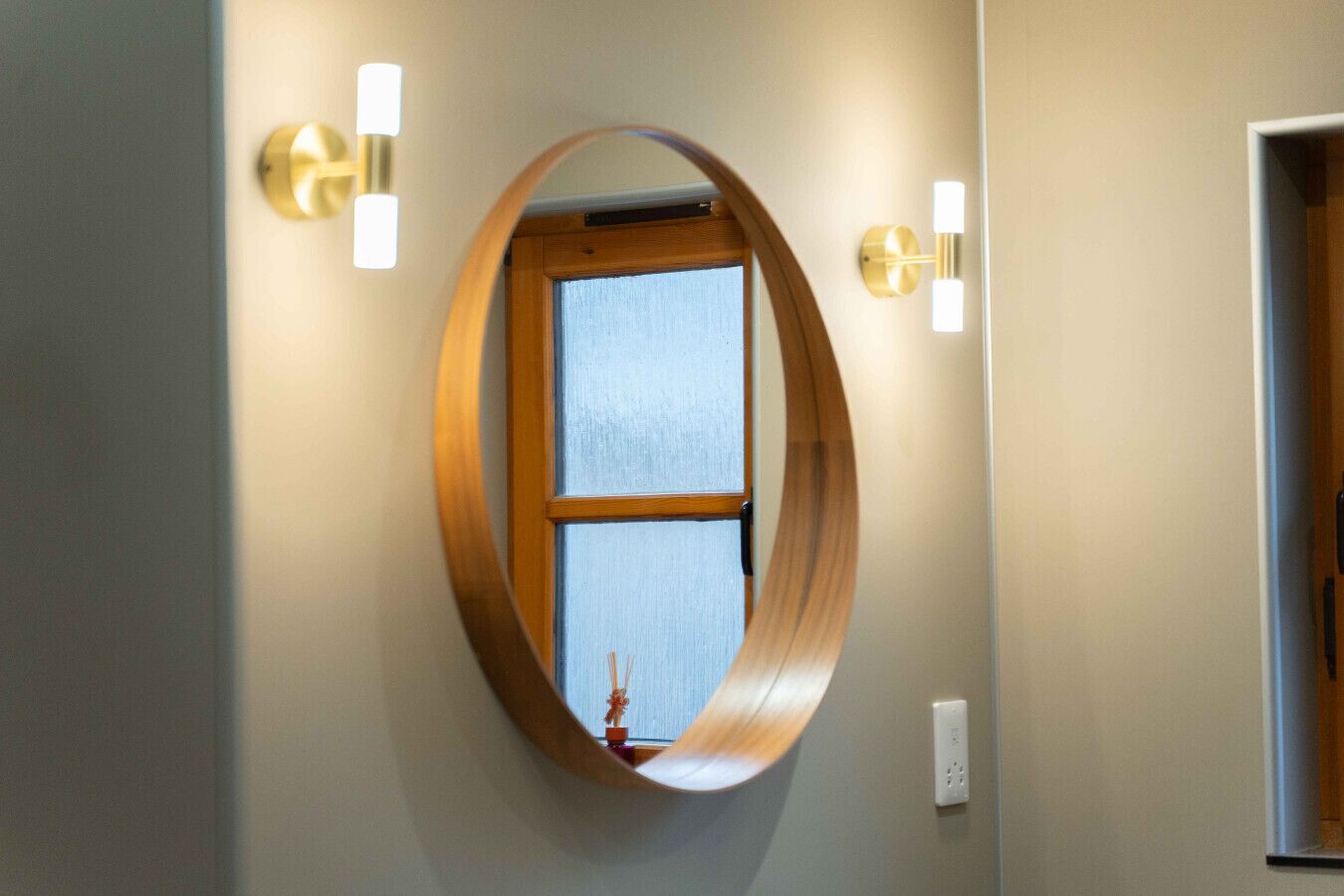 A round mirror with a wooden frame reflecting a window and a wall-mounted light fixture.