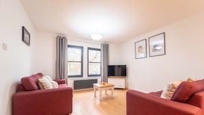 Una sala de estar con dos sofás rojos, una mesita de madera, un televisor sobre un mueble blanco y ventanas amplias con cortinas.