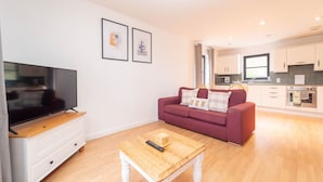 Una sala moderna con un televisor de pantalla plana, una mesa de centro de madera y un sofá rojo con cojines estampados. Se ve una cocina con armarios blancos y un microondas al fondo.