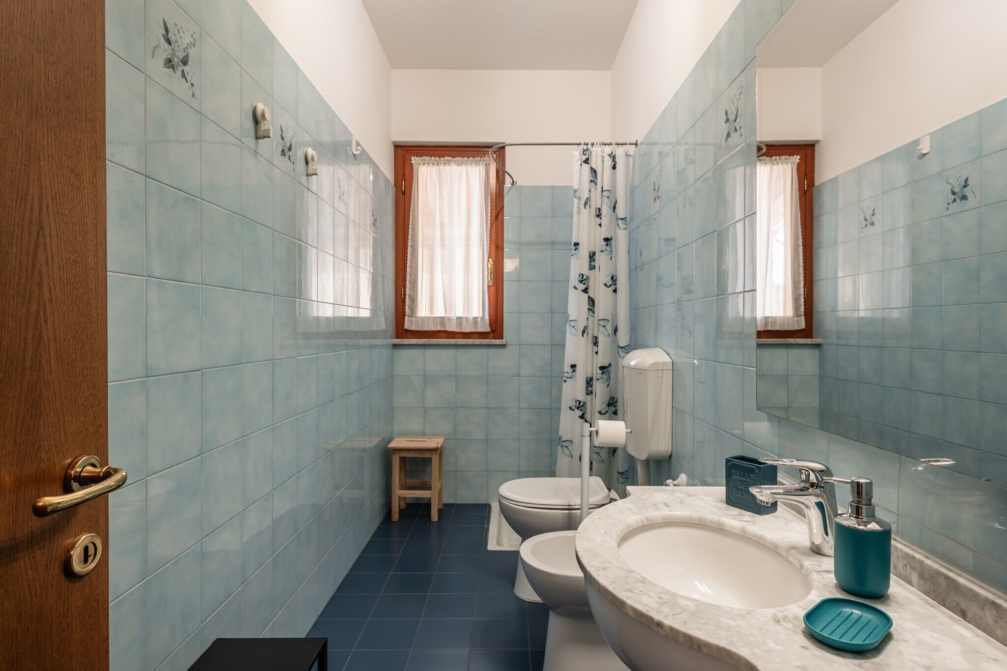 Un baño con azulejos azules, una puerta de madera, un inodoro, un lavabo y un taburete pequeño.