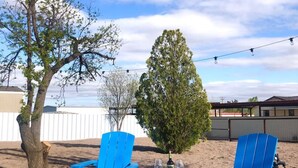 Two blue Adirondack chairs, a small table, and a bottle of wine on a sandy ground with trees and a cloudy sky in the background. - The Art House in Marfa (Marfa)