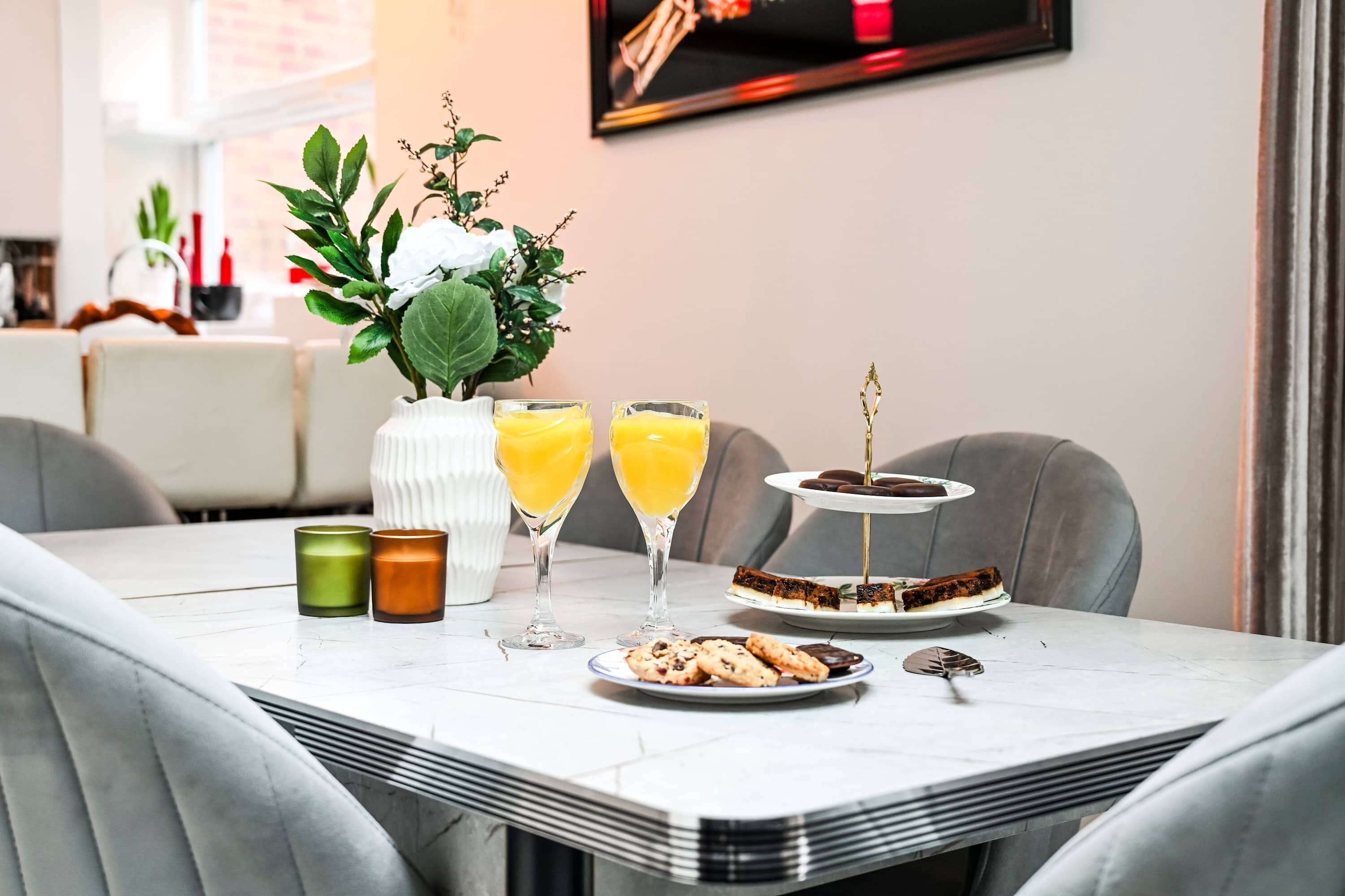 Une table à manger avec une assiette à dessert, deux verres de jus d’orange et un petit vase contenant des feuilles vertes.