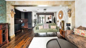A spacious living area with a green sofa, a brown sofa, and a dining table. There is a kitchen area in the background with dark cabinets and a countertop.